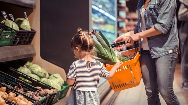 Kleines Kind steht vor Gemüseregal mit Mama und Einkaufskorb