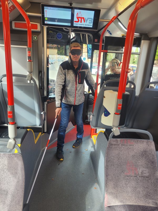 Sehender Mann mit Augenbinde und Pendelstock versucht in einen Bus einzusteigen