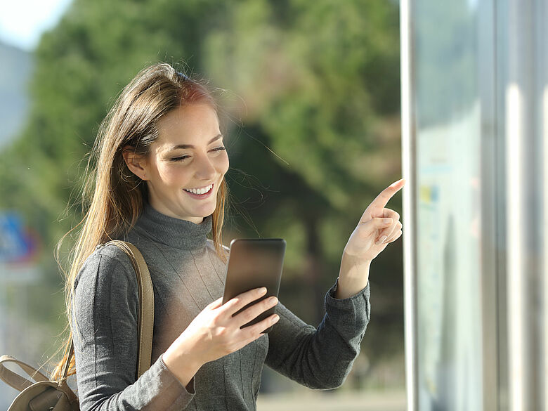 Junge Frau an Bushaltestelle schaut auf den Fahrplan und ins Handy