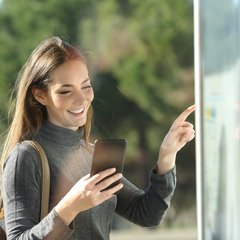 Junge Frau an Bushaltestelle schaut auf den Fahrplan und ins Handy